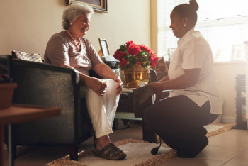 Senior women sitting on a chair at home with female caregiver holding blood pressure gauge.